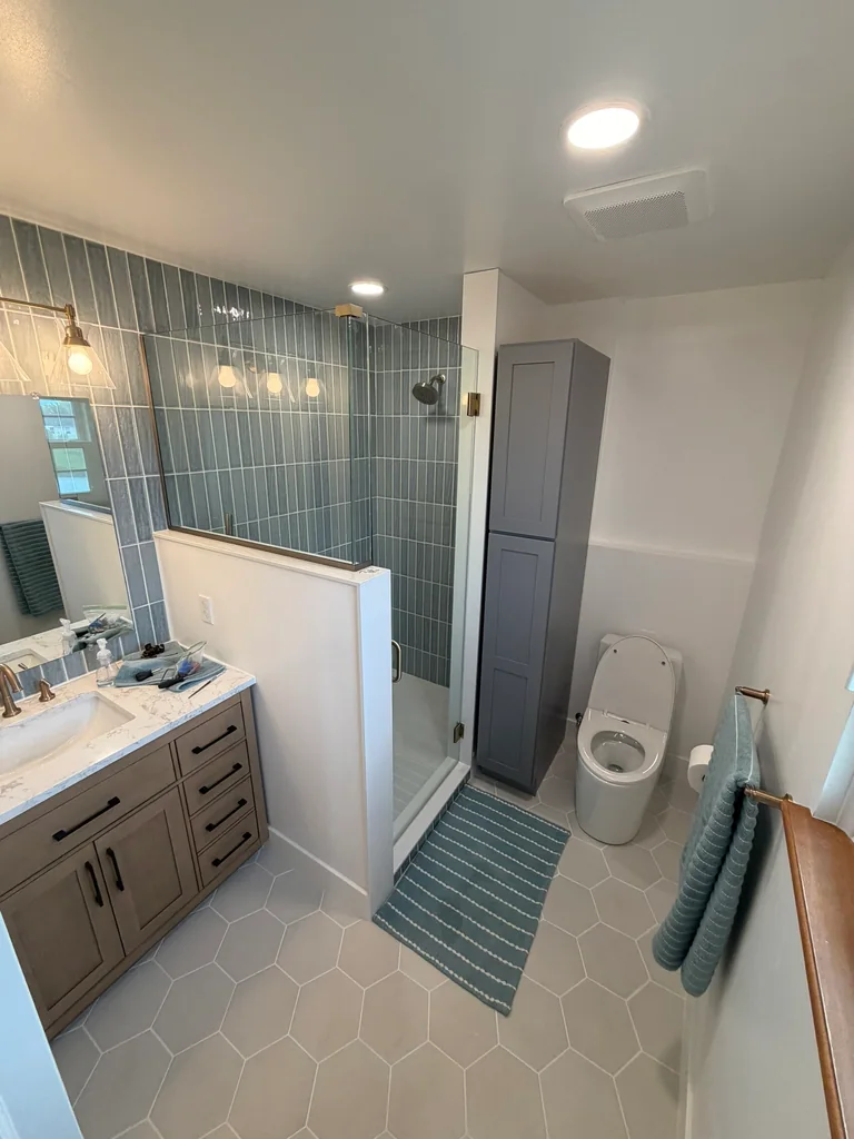 Wood-tone bathroom vanity beside a glass shower, tall gray storage cabinet, and light hexagon floor tile.