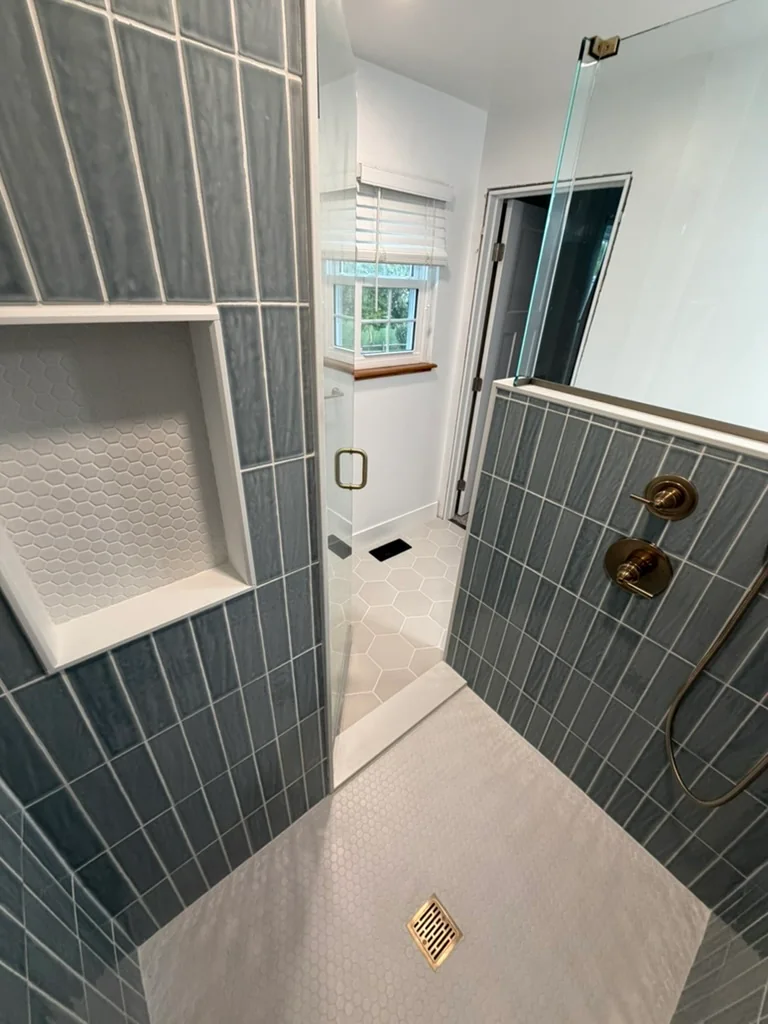Walk-in shower with vertical blue wall tile, a recessed niche, white hexagon mosaic floor, and brass-tone fixtures.